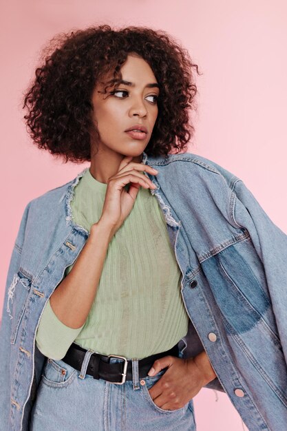 Femme à la peau foncée en tenue de jeans et t-shirt vert posant pensivement sur fond rose Portrait de dame en veste en jean et jupe sur fond isolé