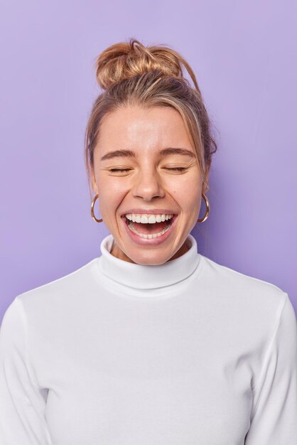 une femme à la peau saine rit joyeusement se sent très heureuse de garder les yeux fermés vêtue d'un col roulé décontracté isolé sur violet. Concept d'émotions et de sentiments positifs des gens