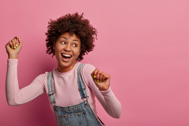 Une femme à la peau sombre et animée insouciante lève les mains, danse joyeusement, regarde du côté droit, aime le temps libre et la liberté, célèbre les bonnes nouvelles, pose contre le mur pastel rose, espace vide de côté