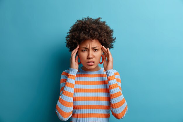 Femme à la peau sombre en détresse touche les tempes et souffre de migraine, de maux de tête douloureux, de vertiges et de sourcils froncés, a besoin d'analgésiques, vêtue d'un pull rayé décontracté, isolé sur un mur bleu