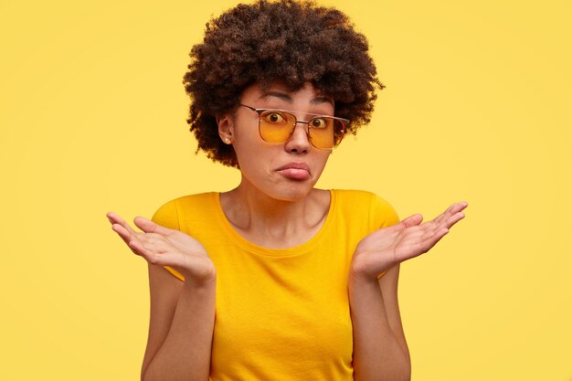 Une femme à la peau sombre incertaine hausse les épaules avec perplexité, vêtue d'un t-shirt jaune décontracté, se sent incertaine