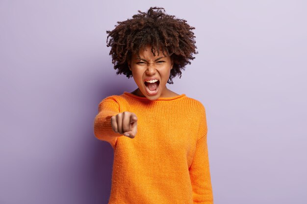 La femme à la peau sombre indignée pointe l'index directement, a une expression agacée, dit que vous êtes coupable, crie avec irritation, se tient contre le mur violet. C'est lui qui m'a offensé