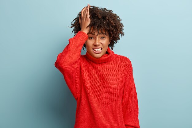 Femme à la peau sombre insatisfaite stressante grimace de douleur, garde la main sur la tête, souffre de maux de tête, a une expression faciale triste impuissante, vêtue d'un pull rouge chaud, isolé sur un mur bleu