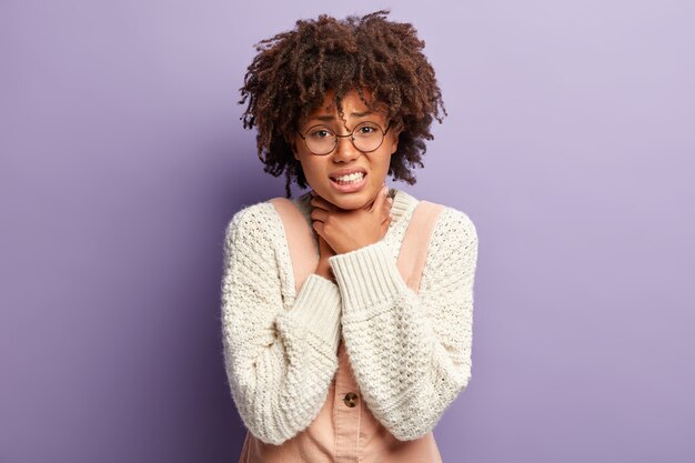 Une femme à la peau sombre et mécontente souffre d'étouffement, garde les deux mains sur la gorge, serre les dents, a les cheveux afro bouclés