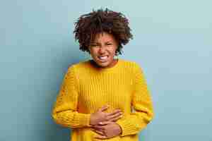 Photo gratuite une femme à la peau sombre et mécontente souffre de maux d'estomac, de crampes menstruelles, garde les mains sur le ventre, porte un pull en tricot jaune, sourit narquoisement, se tient sur un fond bleu.