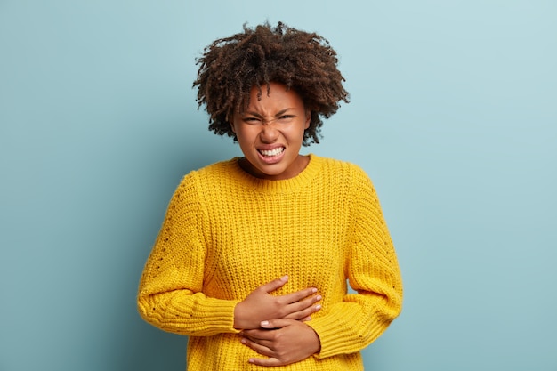 Une femme à la peau sombre et mécontente souffre de maux d'estomac, de crampes menstruelles, garde les mains sur le ventre, porte un pull en tricot jaune, sourit narquoisement, se tient sur un fond bleu.