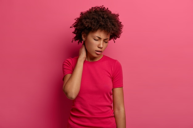 Photo gratuite une femme à la peau sombre mécontente touche le cou, ressent une douleur dans le dos après être tombée d'un escalier, souffre de sentiments douloureux à la colonne vertébrale, incline la tête et ferme les yeux, porte un t-shirt décontracté, isolé sur un mur rose