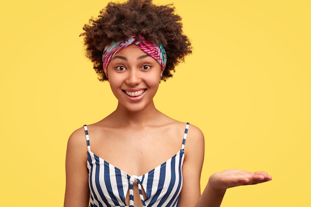 Photo gratuite femme à la peau sombre positive avec une coiffure afro, tient la paume levée, sourit largement, fait semblant de tenir un nouvel article, porte un haut rayé