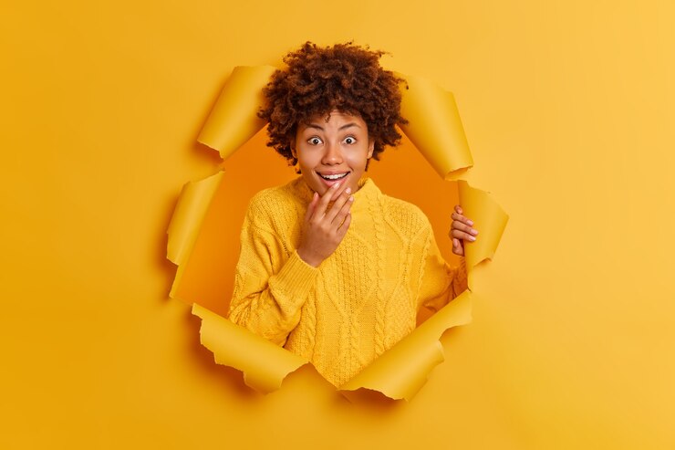 Photo gratuit une femme à la peau sombre positive a une réaction surprise heureuse à travers le trou déchiré de fond de papier
