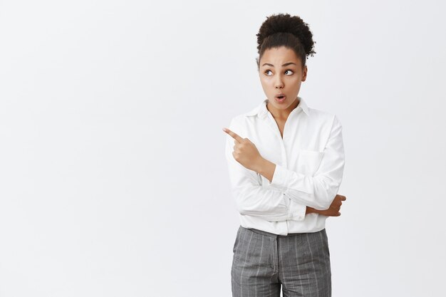 Femme à la peau sombre surprise et étonnée pointant le coin supérieur gauche avec une expression impressionnée