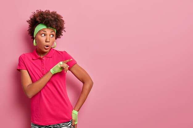 Une femme à la peau sombre surprise et impressionnée regarde avec émerveillement, panique et pointe du doigt, laissée sans voix sur un événement choquant qui s'est produit, porte un bandeau vert, un t-shirt et des gants de sport