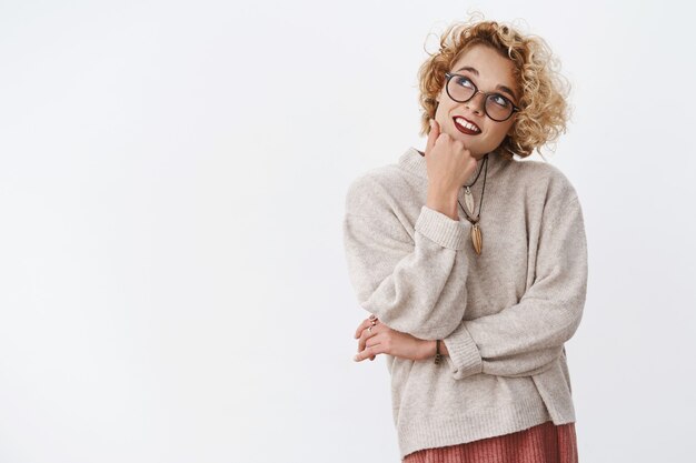 femme pensant quel ordre pour le déjeuner rappelant les restaurants locaux, tenant la main sur le menton et regardant le coin supérieur gauche rêveur et réfléchi avec un léger sourire mignon portant des lunettes et un pull hipster.