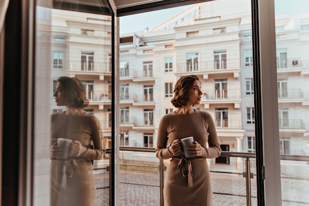 Femme pensive en robe brune buvant un cappuccino. Portrait d'incroyable jolie fille avec une tasse de café debout près du balcon.