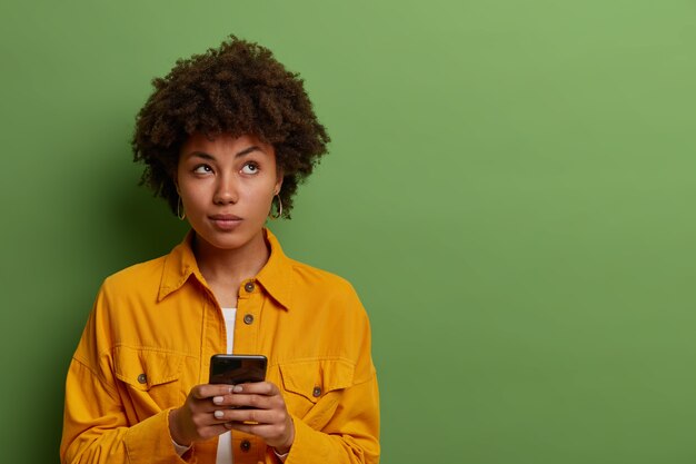 Femme pensive utilise une application moderne sur smartphone, pense au contenu du message, concentré au-dessus, reste en contact avec les technologies modernes, vêtue de vêtements à la mode, espace vide sur le mur végétal