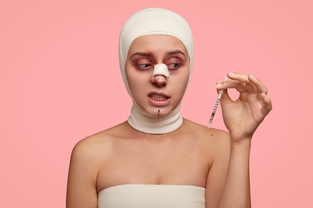 Femme perplexe avec des lignes pointillées sur le visage, a des procédures anti-âge, tient une seringue avec du sérum, enveloppée dans un bandage médical