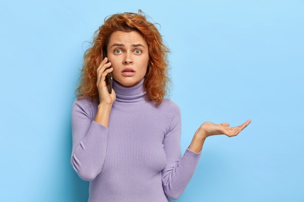 Une femme perplexe et mécontente a des cheveux roux naturels, parle via un téléphone portable, raconte une situation confuse qui lui est arrivée
