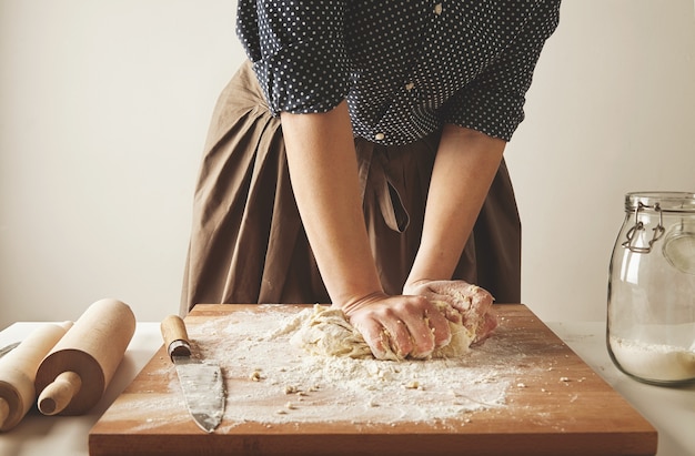 Femme pétrit la pâte pour pâtes sur planche de bois près de deux rouleaux à pâtisserie et pot de farine