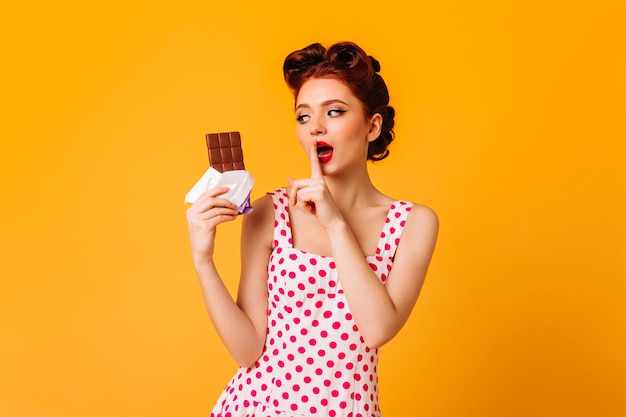 Femme pin-up sensuelle, manger du chocolat sur l'espace jaune. Fille de gingembre en robe à pois touchant les lèvres avec le doigt.