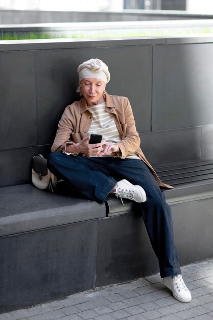 Femme plus âgée à l'extérieur de la ville assise sur un banc et utilisant un smartphone