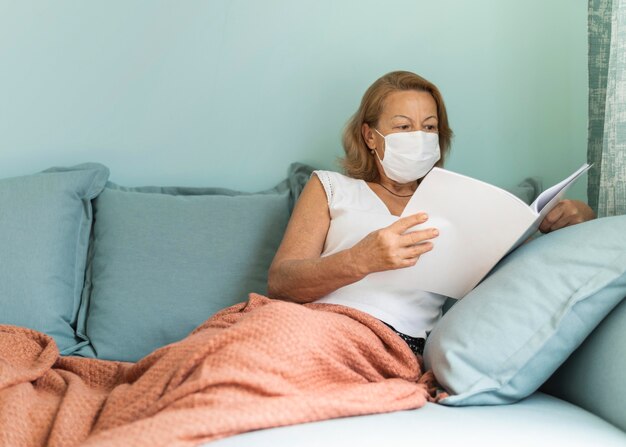 Femme plus âgée avec masque médical à la maison pendant la pandémie en lisant un livre