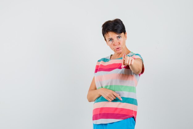 Femme pointant vers la caméra en t-shirt rayé, pantalon et à la vue de face, hésitante.