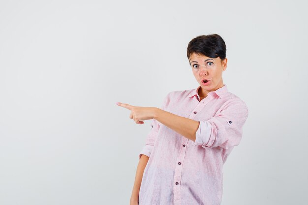 Femme pointant vers le côté gauche en chemise rose et à la surprise, vue de face.