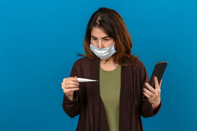 Femme portant un cardigan marron en masque de protection médicale holding smartphone regardant thermomètre numérique en main appelant à quelqu'un à la nervosité sur mur bleu isolé