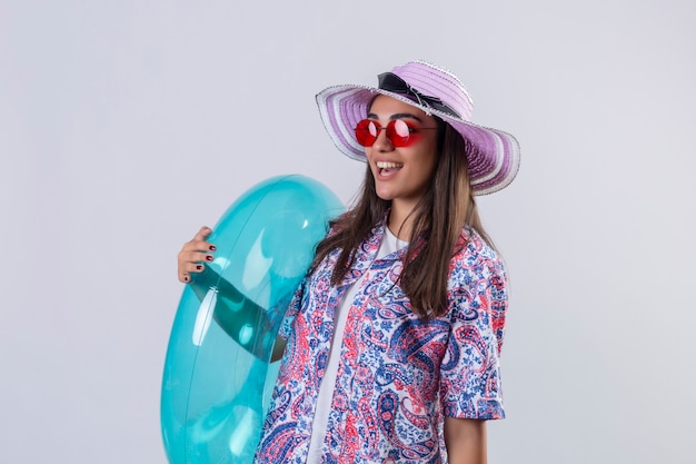 Femme portant un chapeau d'été et des lunettes de soleil rouges tenant un anneau gonflable à la joyeuse positive et heureuse souriant joyeusement debout sur blanc isolé