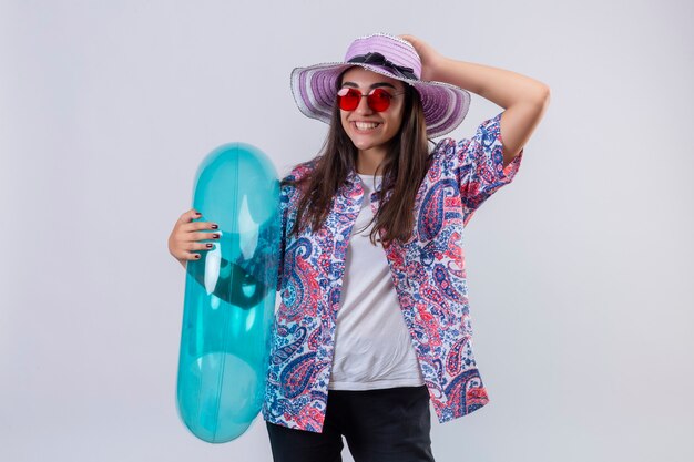 Femme portant un chapeau d'été et des lunettes de soleil rouges tenant un anneau gonflable positif et heureux souriant joyeusement touchant son chapeau debout sur blanc