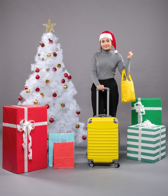 Femme portant un chapeau de père Noël avec des bagages à côté de l'arbre de Noël