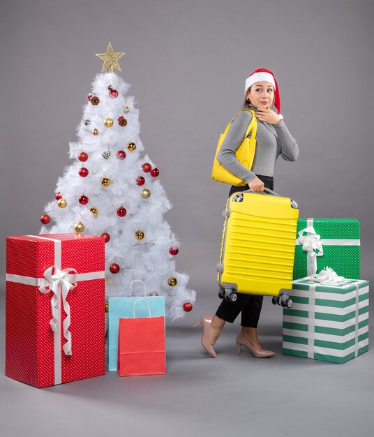 Femme portant un chapeau de père Noël avec des bagages à côté de l'arbre de Noël