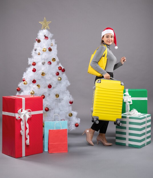 Femme portant un chapeau de père Noël avec des bagages à côté de l'arbre de Noël