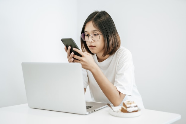 Une femme portant une chemise blanche, jouant avec un smartphone et ayant un ordinateur portable.