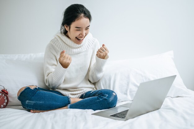 Une femme portant une chemise blanche sur le lit et jouant joyeusement avec un ordinateur portable.