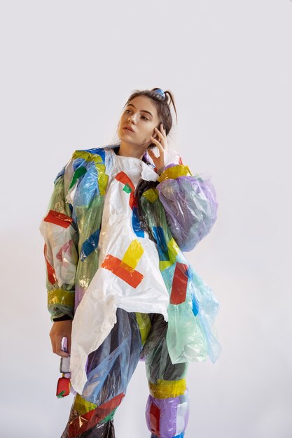 Femme portant du plastique sur un mur blanc. Modèle féminin en vêtements et chaussures faites de déchets. Mode, style, recyclage, concept écologique et environnemental. Trop de pollution, on en mange et on en prend.