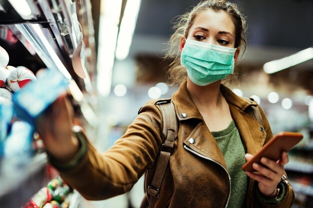 Femme portant un masque de protection lors de l'utilisation d'un téléphone portable et de l'achat de nourriture à l'épicerie pendant l'épidémie de virus