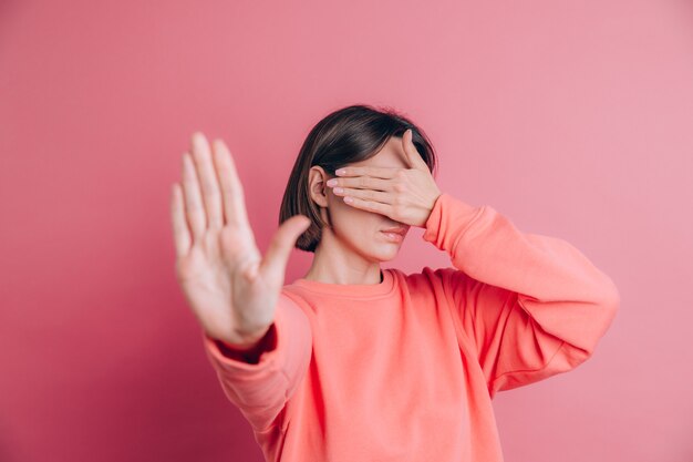 Femme portant un pull décontracté sur fond couvrant les yeux avec les mains et faisant un geste d'arrêt avec une expression de tristesse et de peur