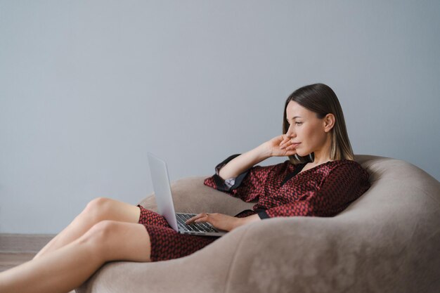 Femme portant une robe de soie à l'aide d'un ordinateur portable à la maison se détendre assis sur un pouf