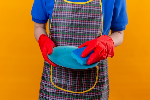 Femme portant un tablier et des gants en caoutchouc tenant la vaisselle dans les mains, plaque de lavage sur mur orange