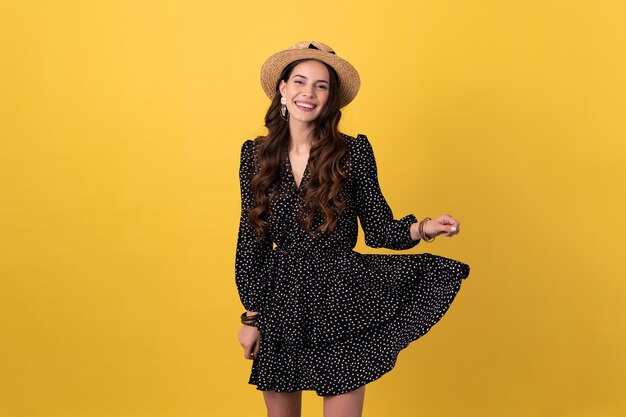 Femme posant isolée sur jaune portant une robe noire à pois et un chapeau de paille boho élégant