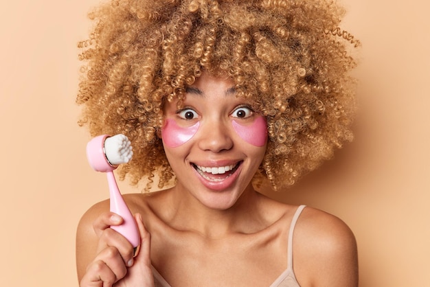 Une femme positive aux cheveux bouclés et aux cheveux bouclés et touffus utilise une brosse spéciale pour un nettoyage en profondeur applique des patchs hydratants sous les yeux prend soin des poses du visage et de la peau à l'intérieur contre un mur beige