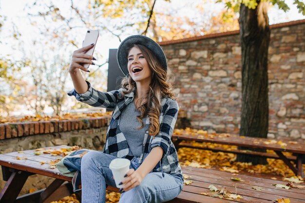 Femme positive aux cheveux brun clair faisant selfie tout en buvant du café dans le parc de l'automne