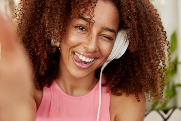 Une femme positive avec une coiffure crépue et touffue montre sa langue, connecte son selfie à un appareil méconnaissable, écoute ses chansons préférées dans une liste de lecture avec des écouteurs, profite du temps libre seul. Ethnicité, mode de vie