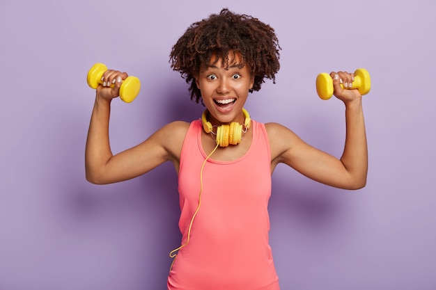 Femme positive avec coupe de cheveux afro, lève les bras avec des haltères, porte des écouteurs jaunes et gilet rose, pose sur le mur violet du studio