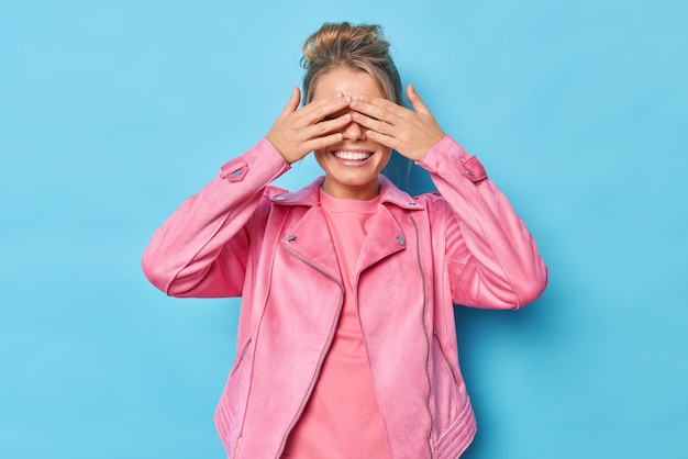 Une femme positive couvre les yeux avec les mains joue à cache-cache des sourires à pleines dents attend la surprise vêtue d'une veste rose décontractée isolée sur fond bleu se sent amusée attend la commande d'ouvrir les yeux