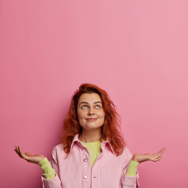 Photo gratuite une femme positive espère que les rêves deviennent réalité, se tient debout avec la paume levée et regarde au-dessus, fait une demande, a les cheveux roux, vêtue de vêtements élégants