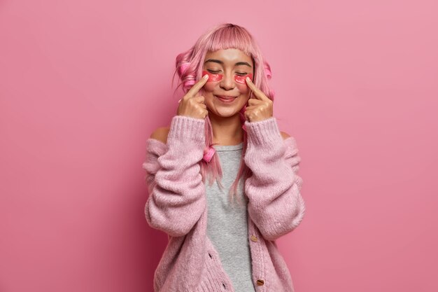 Une femme positive et heureuse a de longs cheveux roses, fait une coiffure frisée avec des bigoudis, indique des patchs de beauté, se tient les yeux fermés porte un pull chaud