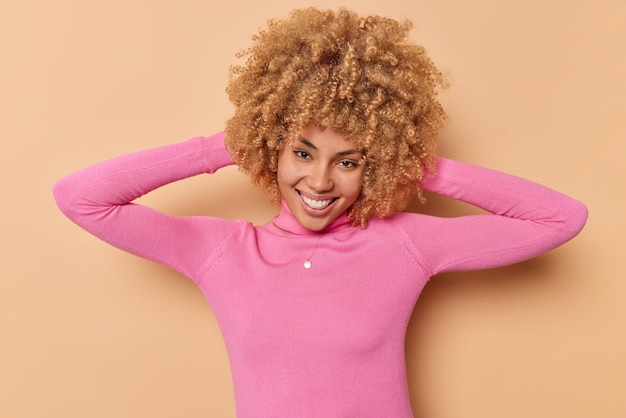 Une femme positive et insouciante garde les mains derrière la tête ressent des sourires joyeux profite joyeusement d'une bonne journée porte un pull à manches longues rose décontracté isolé sur fond beige. Heureux belle adolescente bouclée