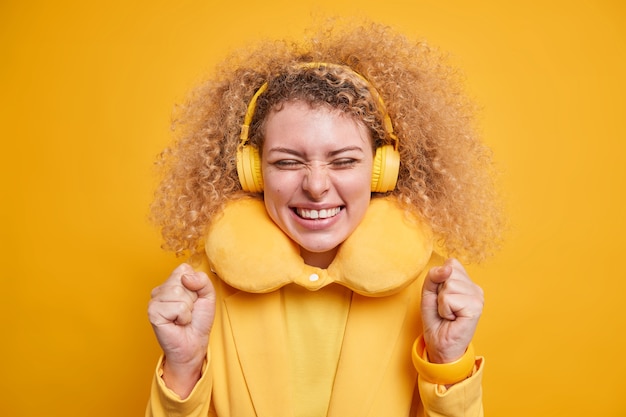 Photo gratuite une femme positive et joyeuse aux cheveux bouclés porte un oreiller confortable autour du cou pour réduire la douleur aime la musique préférée utilise des écouteurs sans fil exprime le bonheur isolé sur un mur jaune