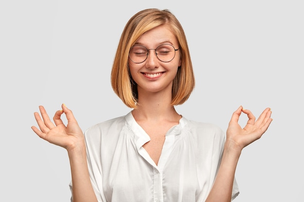 Une femme positive joyeuse fait un geste correct avec les deux mains, a une coupe de cheveux à la mode, ferme les yeux, vêtue d'une chemise blanche